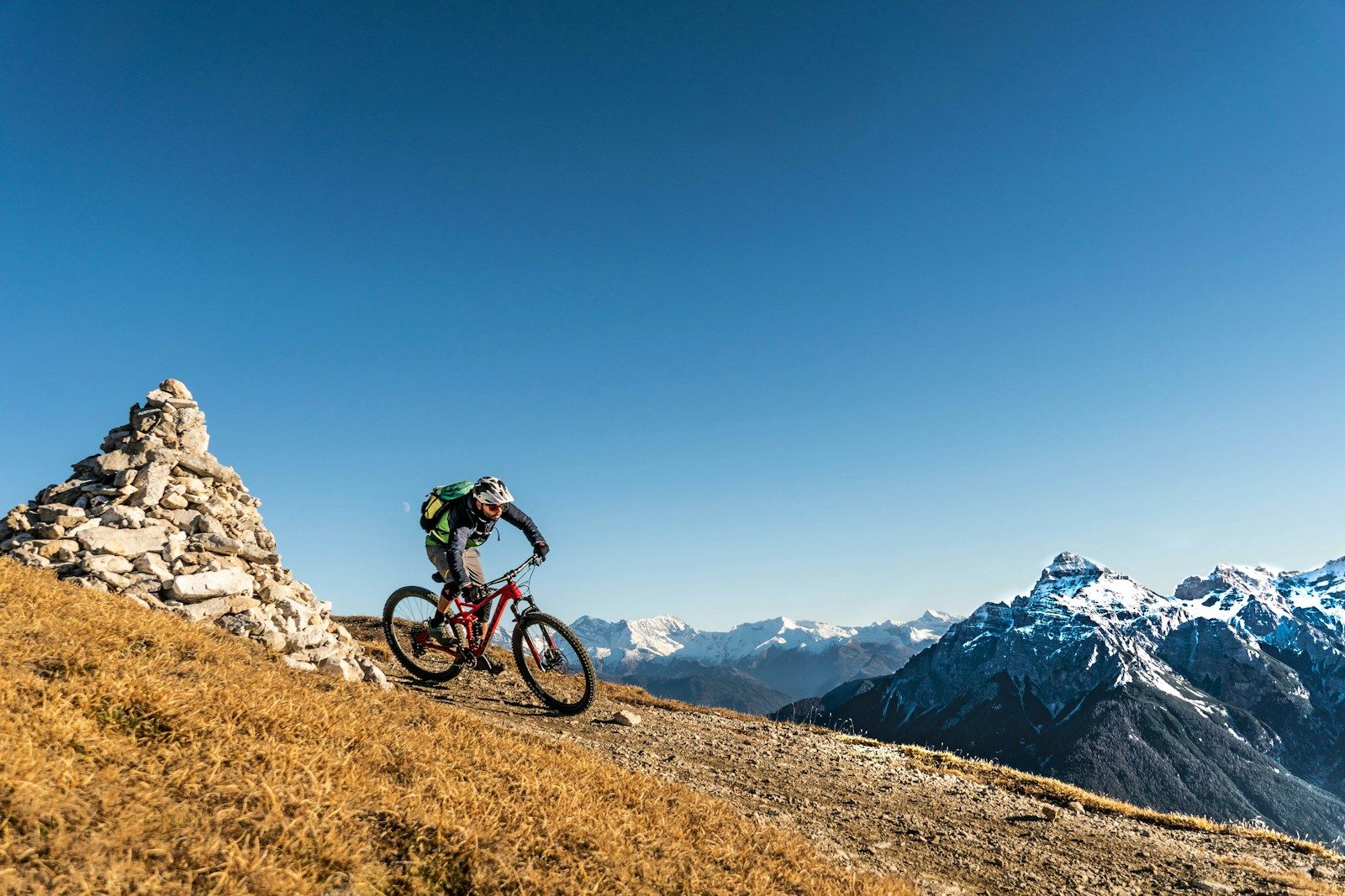 A mountain biker riding past a stone cairn on a high ridge with snowy peaks behind