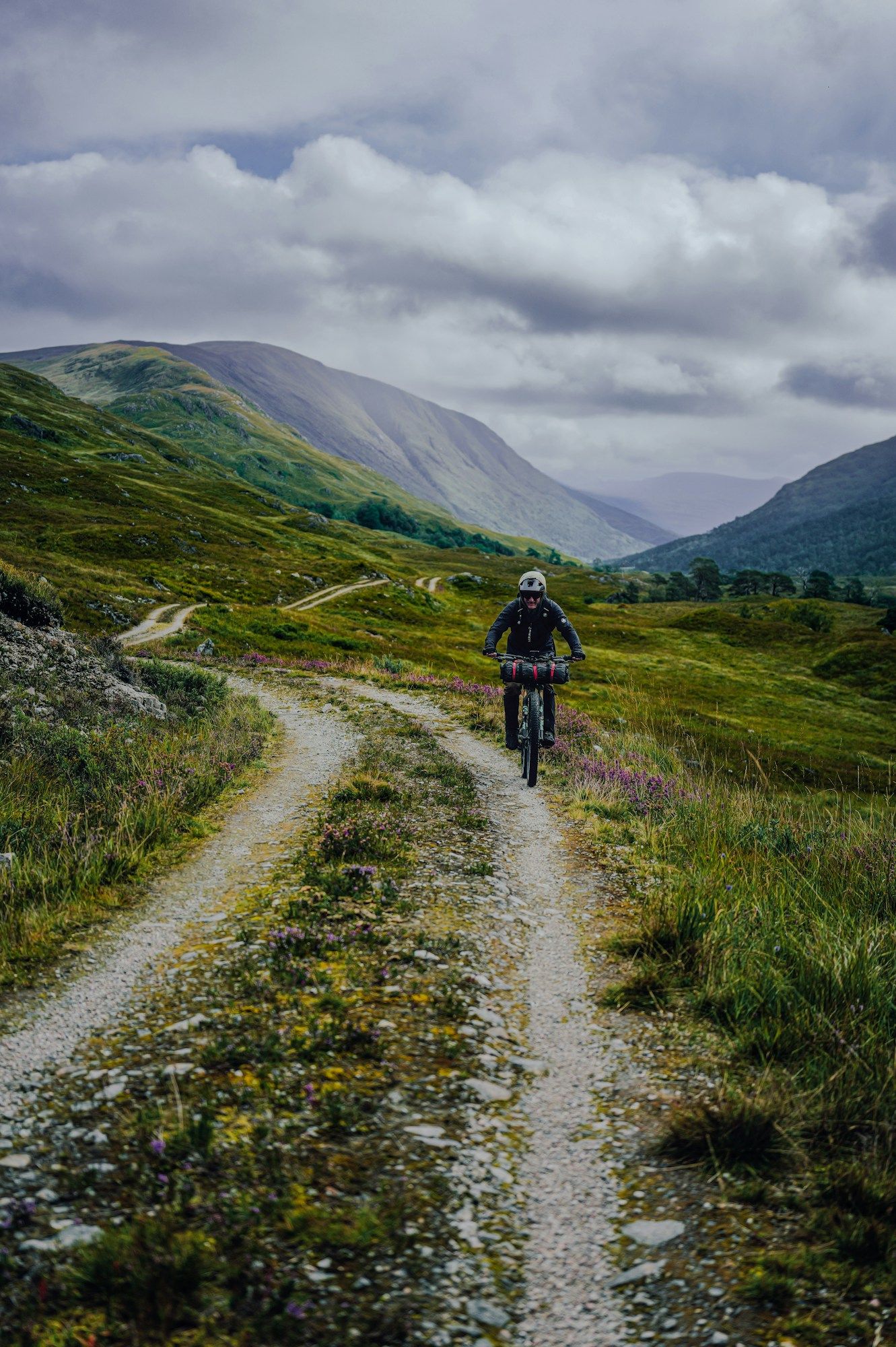 A cyclist on a gravel track running up a green mountain valley under a cloudy sky