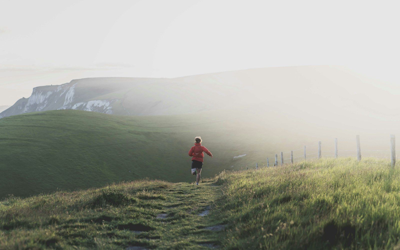 Una corredora con chaqueta roja en un sendero de ladera verde entre la niebla