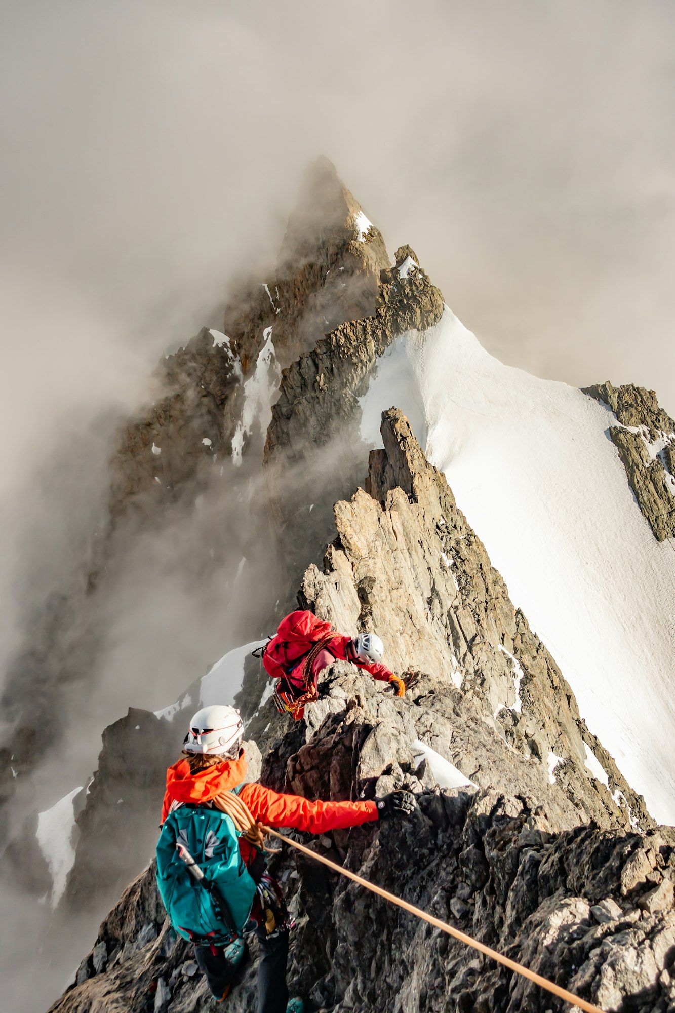 Dos escaladores encordados con casco avanzan por una cresta rocosa hacia una cima nevada entre las nubes