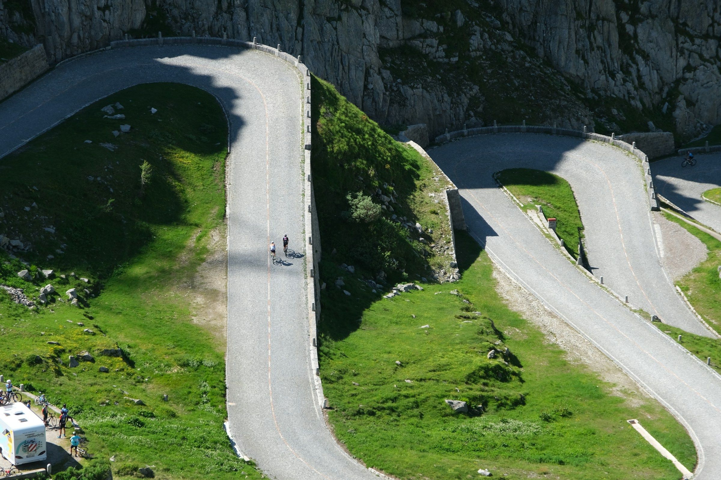 Two cyclists climbing a cobbled mountain road that doubles back on itself through green alpine slopes