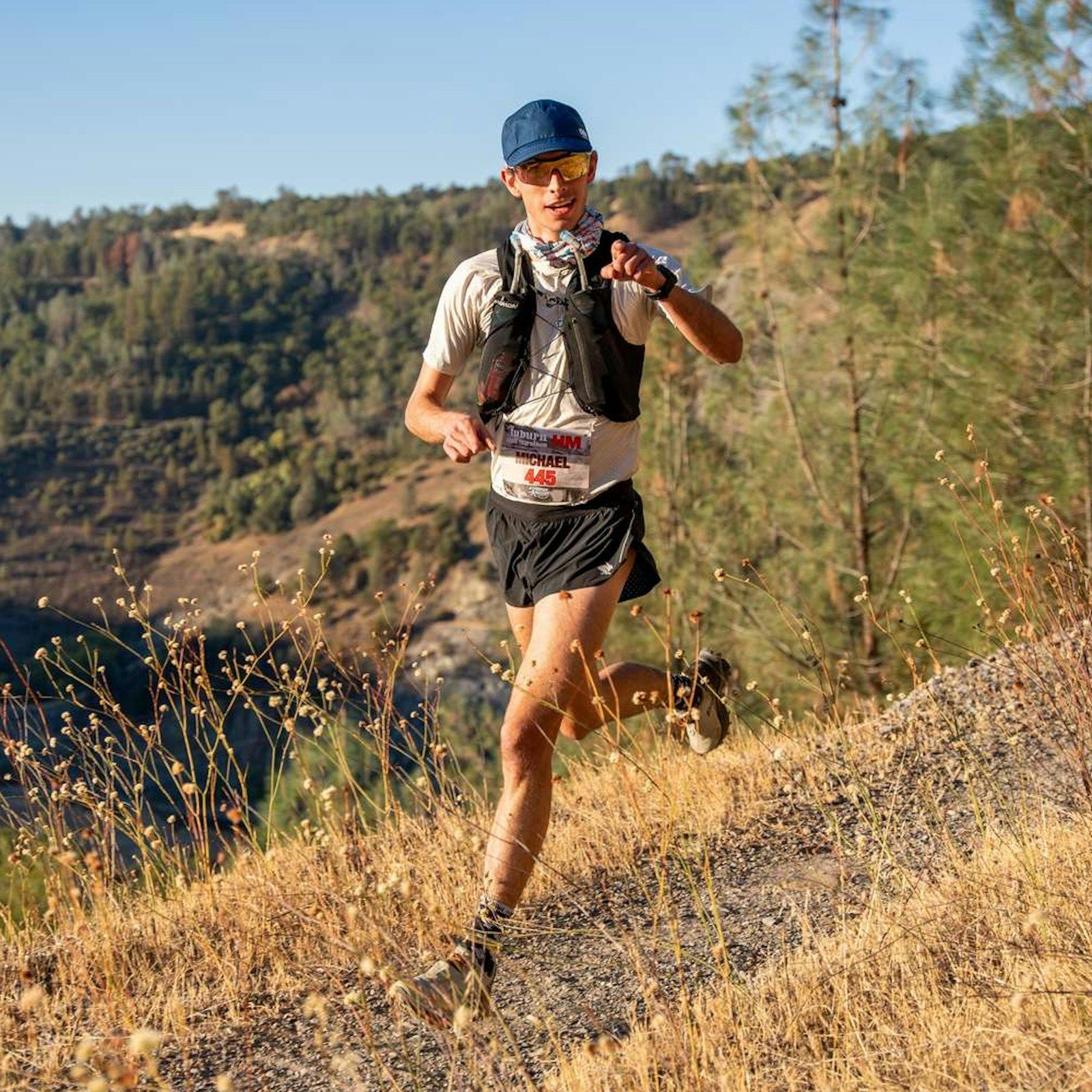 A trail runner with a race bib and running vest on a dry hillside path above a forested canyon